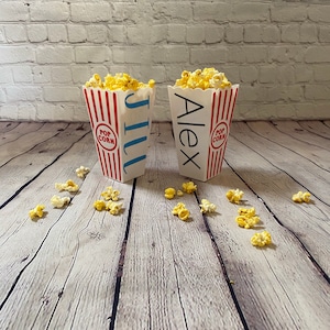 May include: Two red and white striped popcorn containers with the names "Jill" and "Alex" printed on them. The containers are filled with popcorn and are sitting on a wooden surface.
