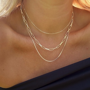 May include: Three gold necklaces layered on a person's neck. The top necklace is a delicate chain, the middle necklace is a thicker chain with oval links, and the bottom necklace is a thin chain with small square links.