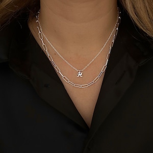 May include: A silver chain necklace with a silver letter 'H' pendant. The necklace is layered over a thicker silver chain.