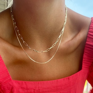 May include: Two gold necklaces layered on a person's neck. The top necklace is a thicker chain with larger, rectangular links. The bottom necklace is a thinner chain with smaller, round links.
