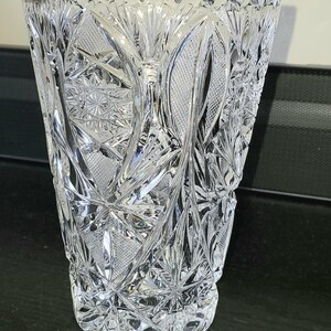 May include: A clear, cut crystal vase with a flared top and intricate geometric patterns. The vase features starburst and diamond designs, creating a sparkling effect. The vase is approximately 20 cm tall.