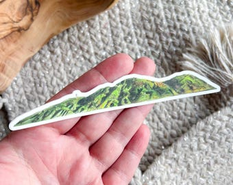 May include: A sticker featuring a mountain range illustration. The design showcases a green and yellow landscape with a white border. The sticker is held in a hand, with a wooden bowl and textured fabric in the background.