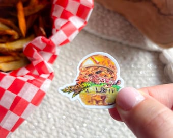 May include: A sticker of a detailed hamburger with a sesame seed bun, lettuce, and a patty. The sticker is held in a hand, with a red and white checkered basket of french fries in the background.