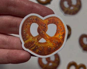 May include: A pretzel sticker with a detailed illustration of a baked pretzel. The sticker has a white border and is held by a hand. Other pretzel stickers are in the background.