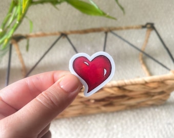 May include: A small, die-cut sticker shaped like a heart. The heart features a deep red gradient with white highlights, giving it a glossy, three-dimensional appearance. It has a thin white border surrounding the entire heart-shaped design.