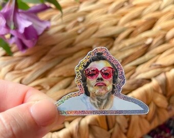 May include: A die-cut holographic sticker featuring a stylized illustration of a person with dark hair wearing bright red heart-shaped sunglasses. The figure is depicted from the chest up, wearing a plain white shirt against a shimmering background.