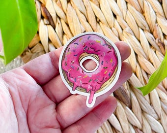 May include: A pink donut with colorful sprinkles and pink icing dripping down the sides. The donut is on a white background.