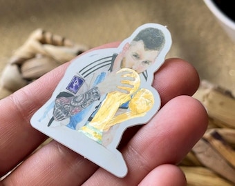 May include: A die-cut sticker featuring a soccer player with dark hair and a beard, wearing a white and light blue striped jersey with a FIFA World Cup patch, kissing a gold-colored trophy. The player has visible arm tattoos and is shown from the chest up.