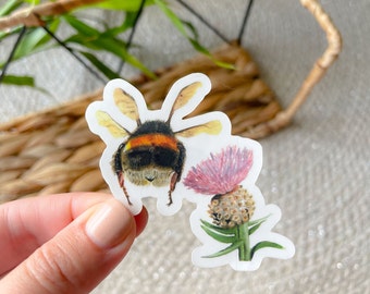 May include: A sticker featuring a detailed illustration of a bumblebee with a black and orange body, and a pink thistle flower. The sticker is held by a hand, with a blurred background of greenery and woven material.