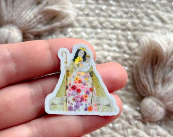 May include: A sticker featuring a religious figure with a gold crown, holding a child. The figure's dress is adorned with colorful flowers. The sticker has a white border and a gold staff.