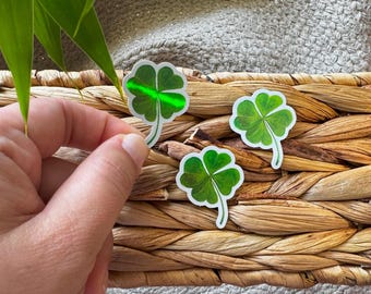 May include: Three green four-leaf clover stickers on a woven wicker surface. The stickers are shiny and have a holographic effect.