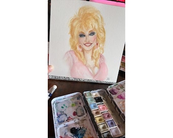 May include: A watercolor portrait painting on textured paper depicting a person with voluminous blonde hair, blue eyes, and pink earrings, wearing a pink top. Below the painting sits a metal watercolor paint tin with various color pans.