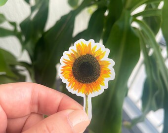 May include: A yellow sunflower sticker with a brown center and green stem. The sticker is on a white background.