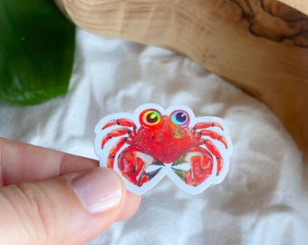May include: A colorful crab sticker with a red body, orange claws, and multi-colored eyes. The sticker is on a white background. The crab sticker is a fun and whimsical design.