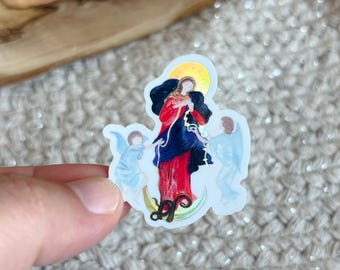 May include: A die-cut sticker featuring a central figure in a red and dark blue robe standing on a crescent moon, flanked by two light blue winged figures. A yellow halo sits above the central figure's head, and a dark serpent coils at the base.