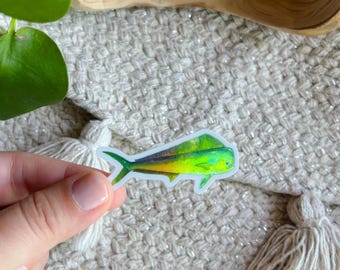 May include: A colorful sticker featuring a mahi-mahi fish. The fish has a green head, yellow and blue body, and a dark blue outline. The sticker is held against a textured, off-white background.