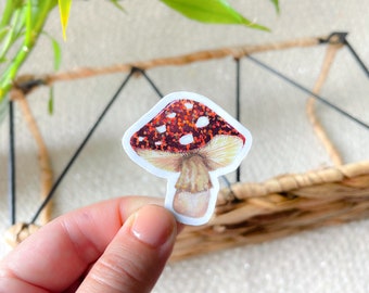 May include: A red and white mushroom sticker with a brown stem. The mushroom has white spots and a glittery texture.