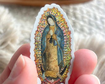May include: A die-cut sticker featuring the Virgin of Guadalupe with a holographic, glittery gold sunburst background. The figure wears a blue mantle over a gold-patterned dress, with hands clasped in prayer, surrounded by a scalloped white border.
