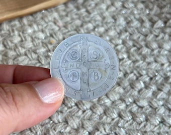 May include: A circular silver-toned Saint Benedict medal featuring a central cross with the letters C S P B and C S M L N D S M D engraved within the quadrants. The medal displays the text PAX at the top and additional Latin lettering around the rim.
