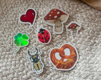 May include: A collection of seven stickers featuring a red heart, a large red and white mushroom, a small red and white mushroom, a green four-leaf clover, a red and black ladybug, a brown stag beetle, and a golden brown salted pretzel.