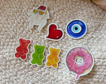May include: A collection of seven stickers featuring a cooked turkey wearing sunglasses and a red hat, a red heart, a blue evil eye, three gummy bears in red, green, and yellow, and a pink frosted donut with colorful sprinkles and glittery accents.