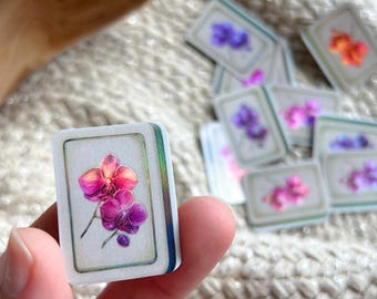 May include: A rectangular tile featuring a vibrant illustration of pink and purple orchid flowers. The tile has a white border with a subtle iridescent sheen along the edges, and it is part of a collection of similar floral tiles.