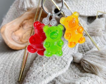 May include: A keychain featuring three gummy bear charms in red, green, and yellow. The charms are attached to a silver metal ring. The background includes a gold-colored decorative element and a soft, textured, light gray surface.