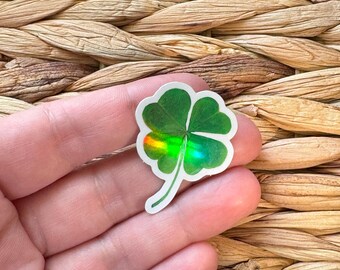 May include: A green four-leaf clover sticker with a holographic finish. The sticker is on a hand.