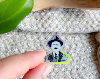 May include: A small, holographic sticker featuring a black and white image of a man in a suit and hat. The sticker has a colorful, iridescent border and is held by a person's fingers. The man has a mustache and is wearing a suit and tie.