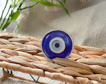 May include: A blue and white evil eye charm with a white center and a blue ring around it. The charm is on a woven wicker surface.