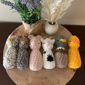 May include: A collection of handmade crochet animal toys, including a chicken, a pig, a cow, a duck, and more. The toys are in various colors, such as white, black, pink, and yellow. They are arranged on a wooden surface with decorative plants.