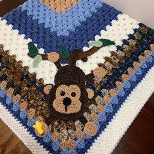 May include: A crocheted baby blanket with a brown monkey holding a yellow banana. The blanket is made with white, brown, blue, and green yarn.