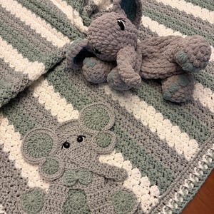 May include: A crochet blanket featuring alternating diagonal stripes in sage green and white. The blanket is adorned with a flat, crocheted elephant appliqué and a three-dimensional, plush, grey crocheted elephant toy with dark eyes.