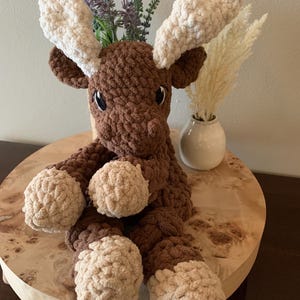 May include: A handmade brown and tan crochet moose plush toy with large white antlers. The moose has a friendly expression and is sitting on a wooden surface. Decorative plants are in the background.