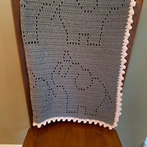 May include: A gray crocheted baby blanket with a pink crocheted border. The blanket is folded and placed on a wooden chair.