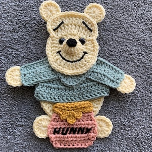 May include: Crocheted Winnie the Pooh applique in cream, green, pink, and gold. The bear has a smiling face with black eyes and a black nose. He wears a green sweater and holds a pink honey pot with the word "HUNNY" stitched on it.