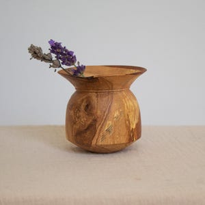 May include: A small, handcrafted wooden vase with a flared top and a rounded body. The vase contains a sprig of dried lavender. The wood grain is visible, displaying natural colours and patterns.