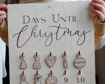 Puede incluir: Un calendario de adviento de madera para Navidad con el texto "DAYS UNTIL Christmas" y los números del 9 al 25. El calendario tiene pequeños adornos de madera colgados de cada número.