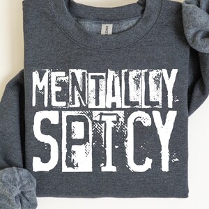 Peut inclure: Sweat-shirt gris fonc&eacute; avec l'inscription "MENTALLY SPICY" en grandes lettres blanches. Le texte "DARK GREY" est imprim&eacute; en dessous. Le sweat-shirt a un col rond et des manches retrouss&eacute;es.