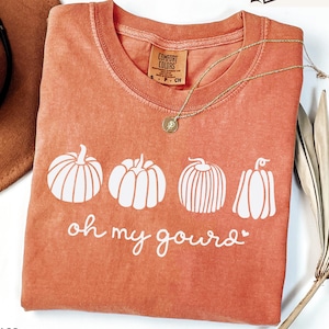 Peut inclure: Un sweat-shirt orange Comfort Colors avec un motif de citrouilles blanches et l'inscription "oh my gourd". Un collier doré avec un pendentif est posé sur le sweat-shirt. Le sweat-shirt est à côté d'un chapeau marron et d'une plante séchée.