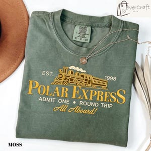 May include: A moss green crew neck t-shirt featuring a gold graphic of a steam locomotive train. The text reads EST. 1998 POLAR EXPRESS ADMIT ONE ROUND TRIP All Aboard! A thin gold chain necklace rests on the collar of the shirt.