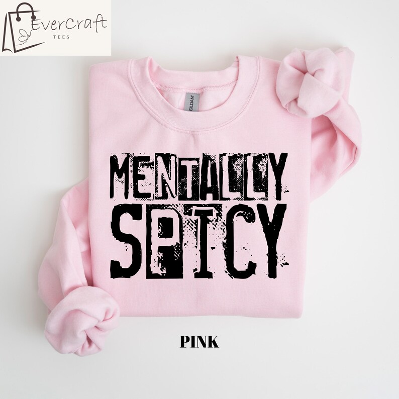 Peut inclure: Un sweat-shirt rose clair avec l'inscription noire "MENTALLY SPICY" dans une police de caract&egrave;res vieillie. Le mot "PINK" est imprim&eacute; en noir en bas. Le logo Evercraft Tees est dans le coin sup&eacute;rieur gauche.