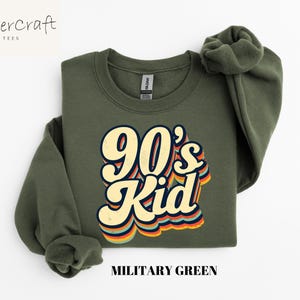 May include: A green sweatshirt with the text "90's Kid" in a retro rainbow font. The sweatshirt is a crew neck style and has a relaxed fit.