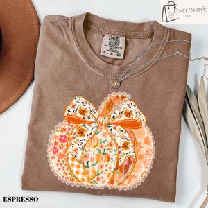 May include: Espresso-colored t-shirt featuring a pumpkin design with a floral bow and lace trim. The pumpkin is filled with various floral patterns and orange accents. A silver necklace with a pendant is draped across the shirt.
