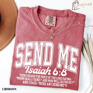 May include: Crimson-colored t-shirt with the text "SEND ME Isaiah 6:8" in white block letters. The shirt also features a quote from Isaiah 6:8. A gold-colored necklace with a pendant rests on the shirt. The shirt is from Comfort Colors.