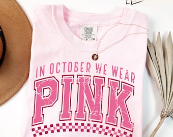 May include: A light pink t-shirt with the text "IN OCTOBER WE WEAR PINK" in a distressed, dark pink collegiate font. The word "PINK" is large and prominent. A rose gold necklace and a brown hat are visible.