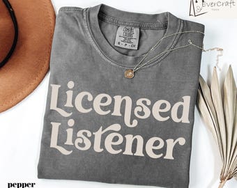 Könnte beinhalten: Ein graues T-Shirt mit den Worten "Licensed Listener" in cremefarbener Retro-Schrift. Das Shirt ist gefaltet und hat ein Comfort Colors-Etikett. Eine silberne Halskette mit einem kleinen Anhänger liegt auf dem Shirt.