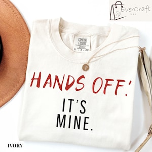May include: An ivory short-sleeved t-shirt featuring the red text "HANDS OFF!" and the black text "IT'S MINE." printed on the front. The shirt has a crew neckline and a Comfort Colors brand label inside the collar, with a small gold pendant necklace.