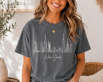 May include: Charcoal grey t-shirt with a white outline of the New York City skyline, including the Statue of Liberty, skyscrapers, and the Brooklyn Bridge. The words "New York" are written in cursive below the skyline.