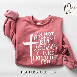 May include: Heather scarlet red sweatshirt with the text "I'M NOT PERFECT BUT JESUS THINKS I'M TO DIE FOR" and a white cross. The long-sleeved top has rolled cuffs and the Evercraft Tees logo in the upper right corner.